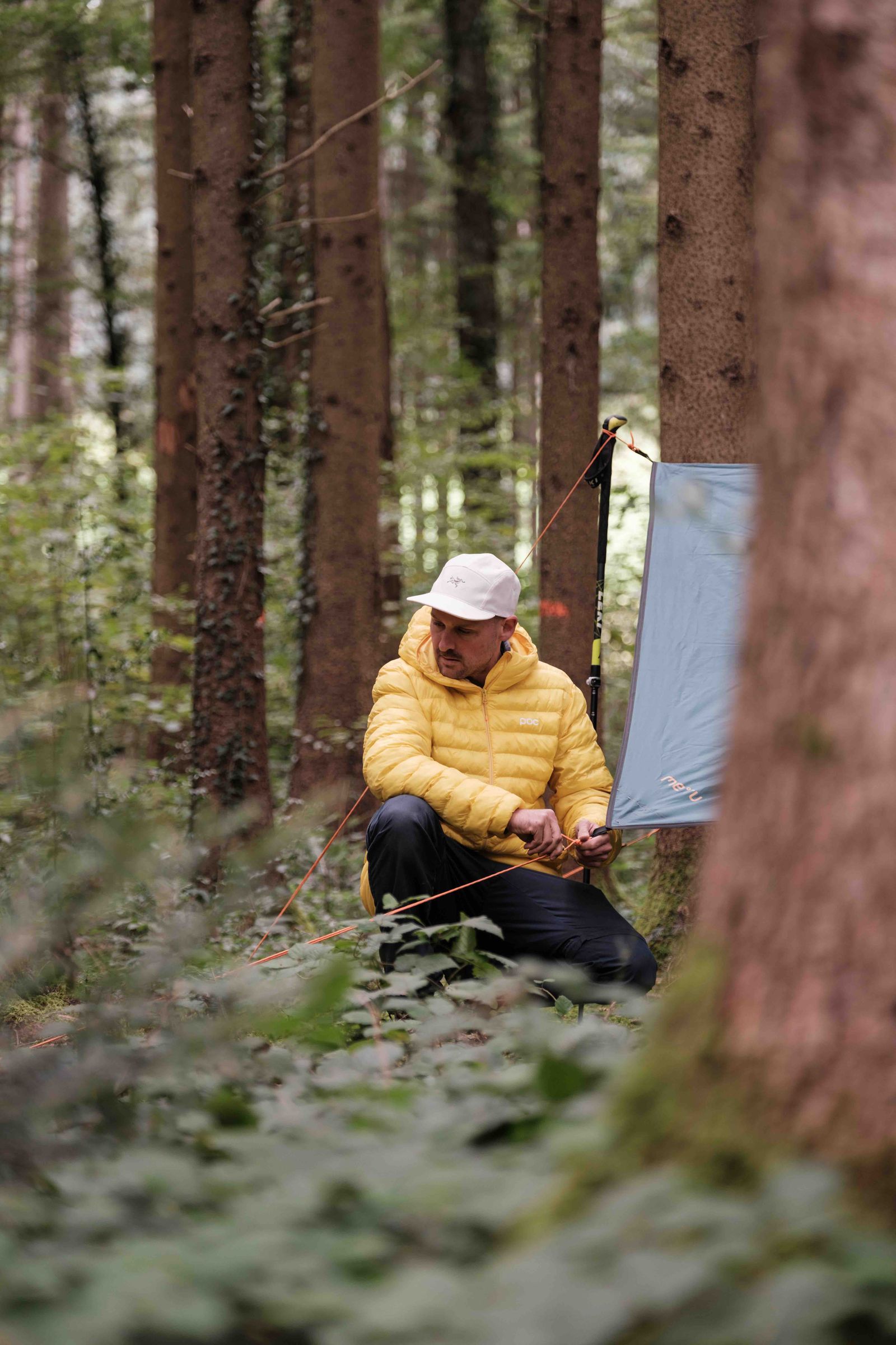 Manuel Seiler, der Outdoorguide in der gelben Daunenjacke mit Cap baut ein Übernachtungscamp im Wald auf mit einer Blache